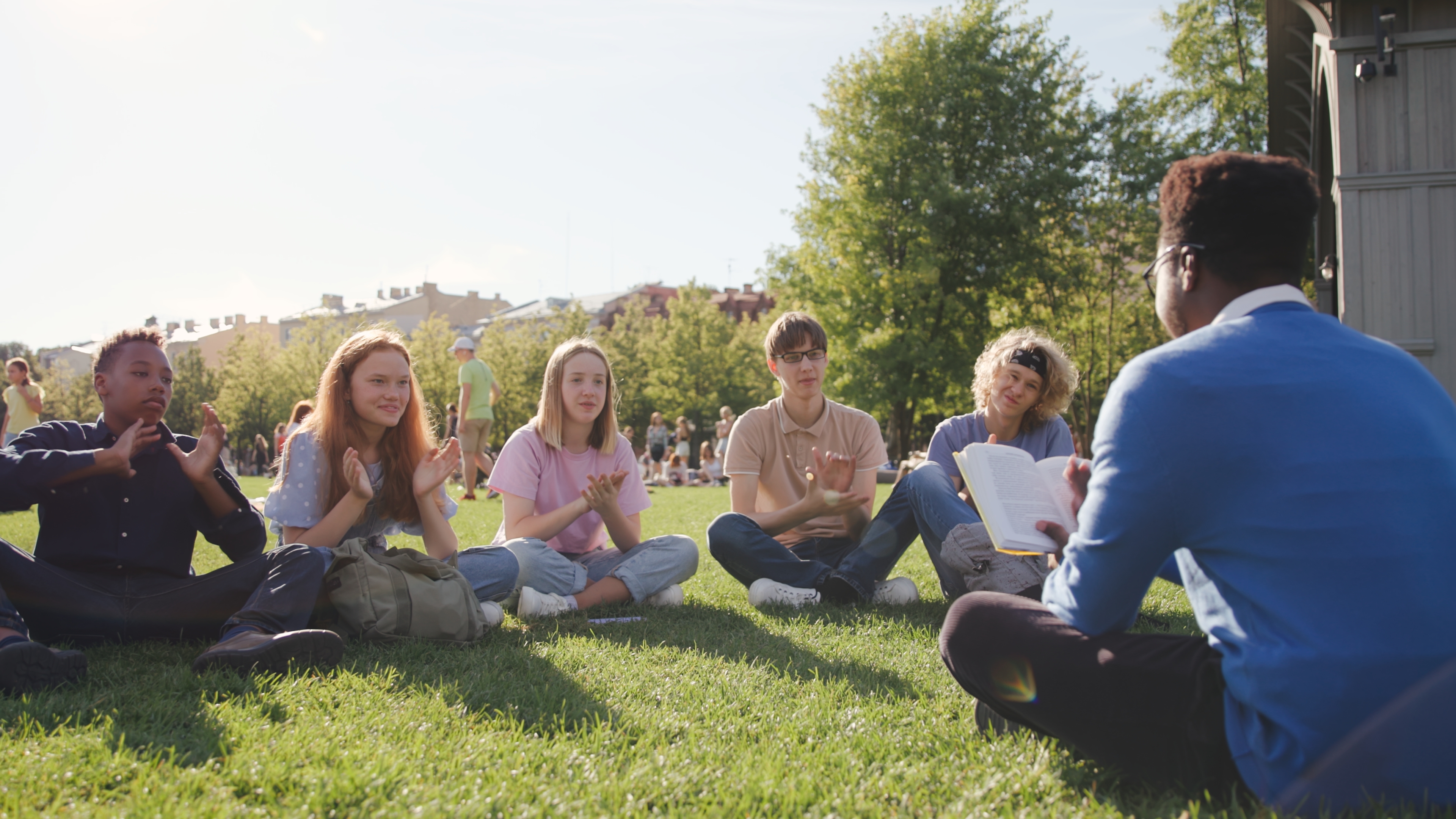 High school students sit on glass and applaud to teacher having outdoor lesson. Back view of African-American professor read to class of diverse students sitting on grass